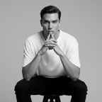 Man sitting on a stool with hands clasped in front of him against a gray background